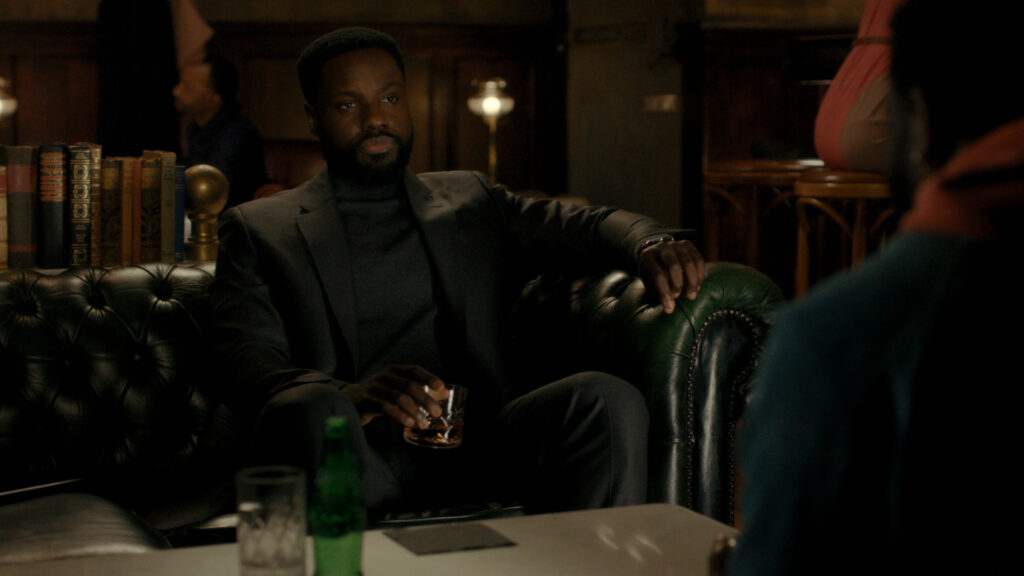 A still from Dark Matter, Leighton sits on a leather sofa drinking Whiskey.
